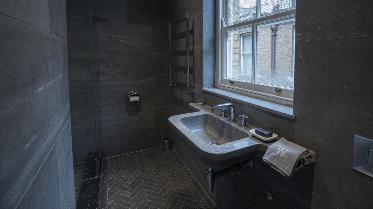 A compact ensuite in a London mansion flat — dark limestone, chrome taps and a lightwell sash