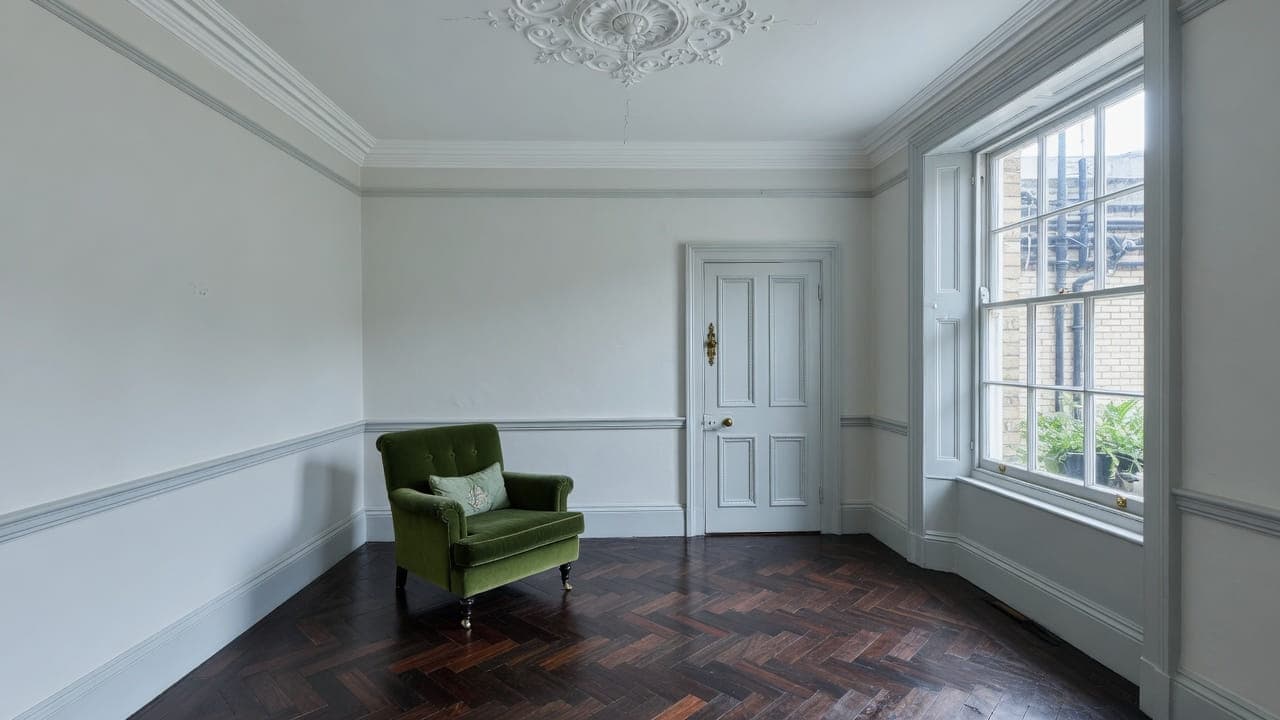 A decorated mansion-flat reception — pale grey walls, parquet, a green armchair and a lightwell sash